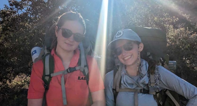 Two young people wearing backpacks smile for a photo in front of greenery. 